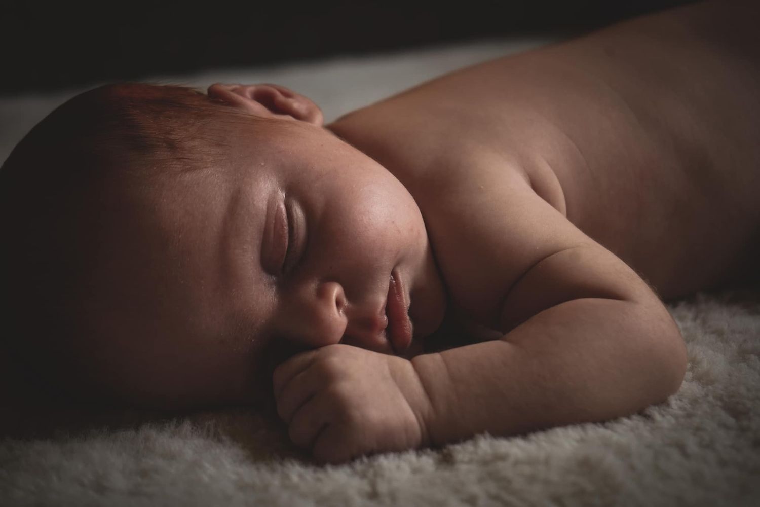 Peaceful newborn baby sleeping on a soft white blanket in a dark, moody setting.