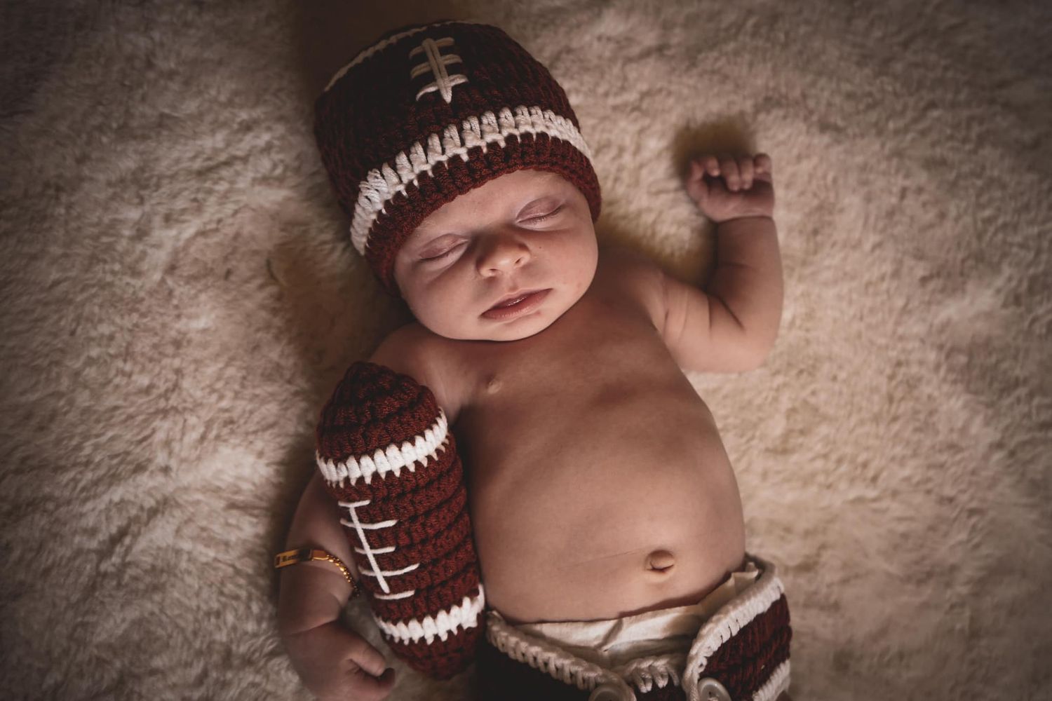 Newborn baby in crocheted football helmet and diaper poses with a football prop.