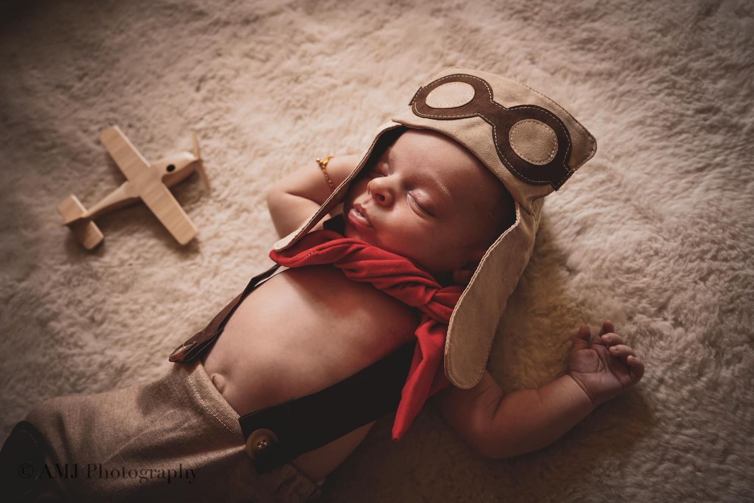 Sleeping newborn dressed as a pilot with goggles and scarf beside a wooden toy airplane.
