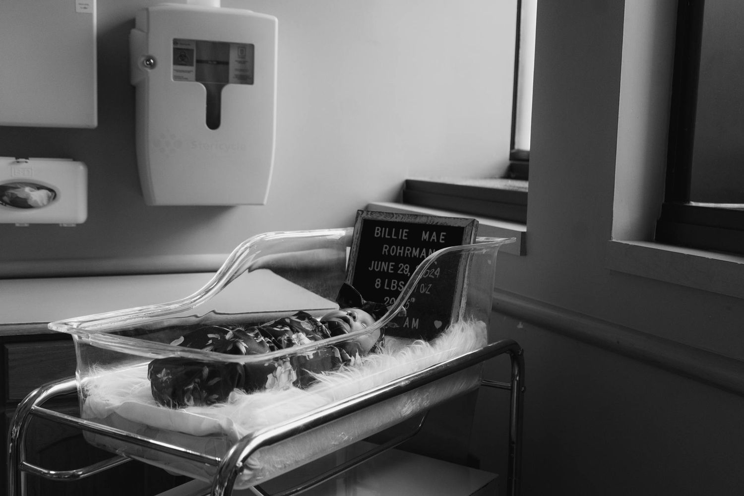 Newborn baby Billie Mae Rohrman resting in hospital bassinet with birth announcement letter board nearby.