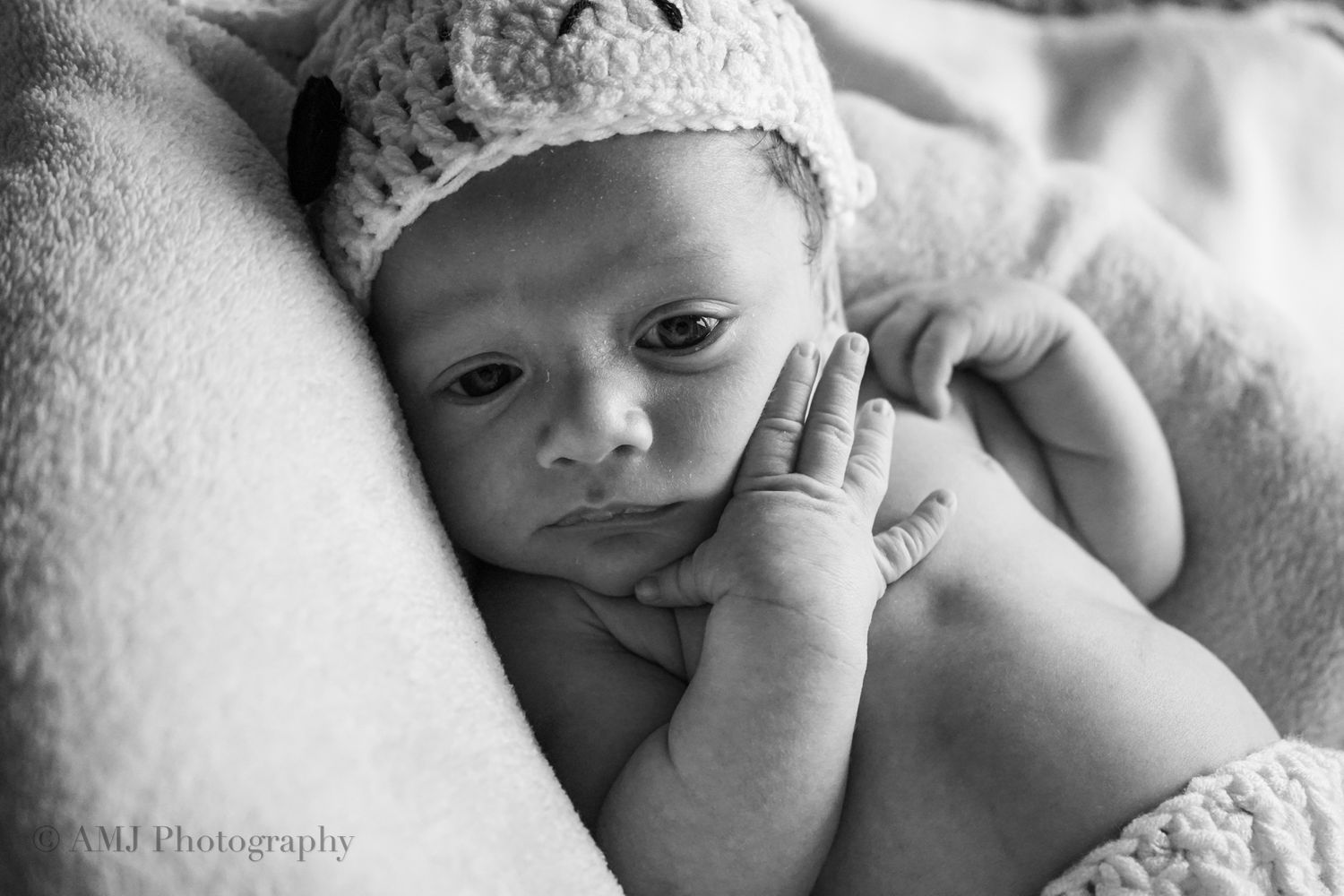 Close-up black and white portrait of alert newborn baby wearing crocheted hat with hand near face.