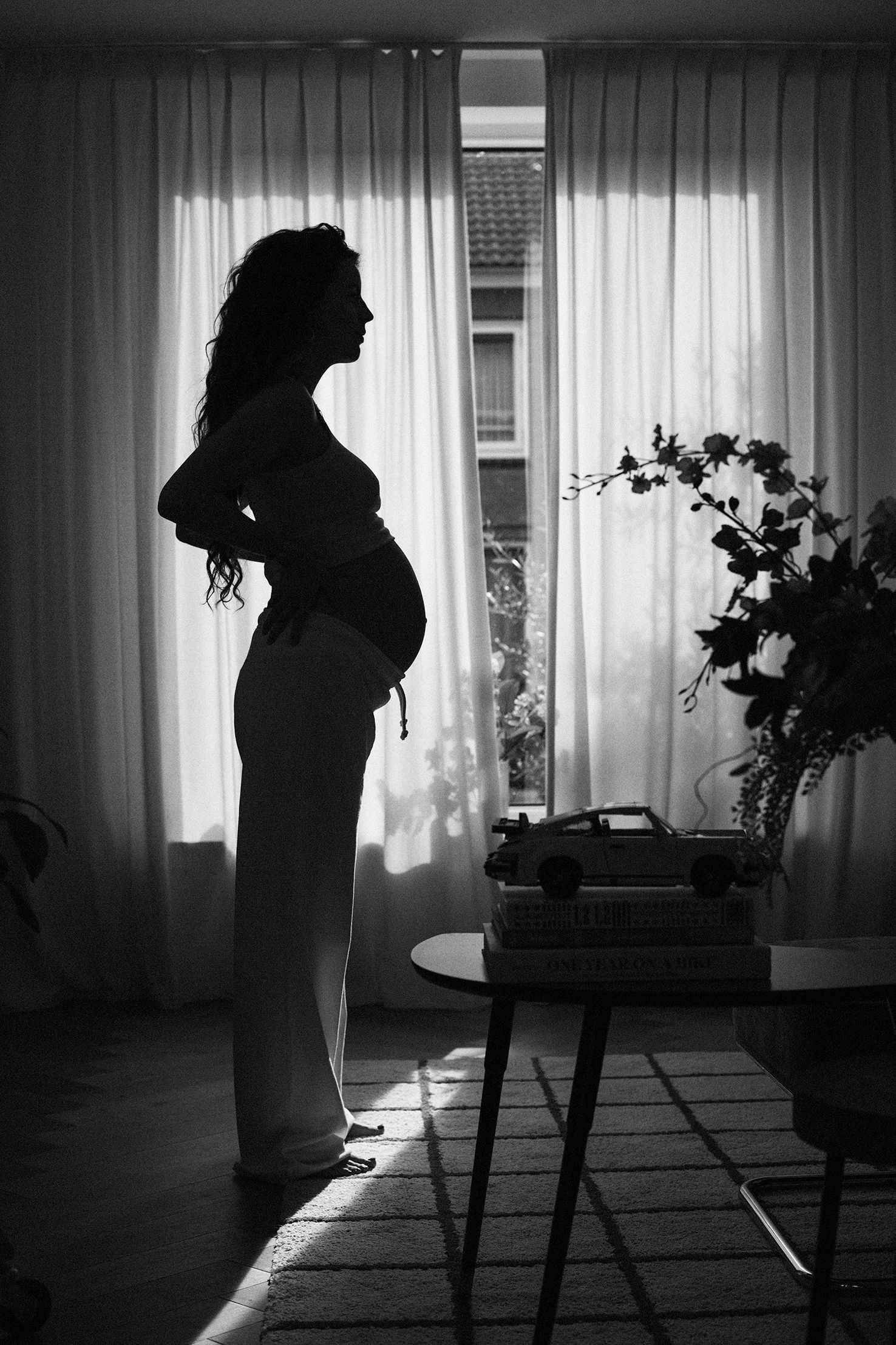 Dramatic black and white silhouette of pregnant figure standing by window with curtains.