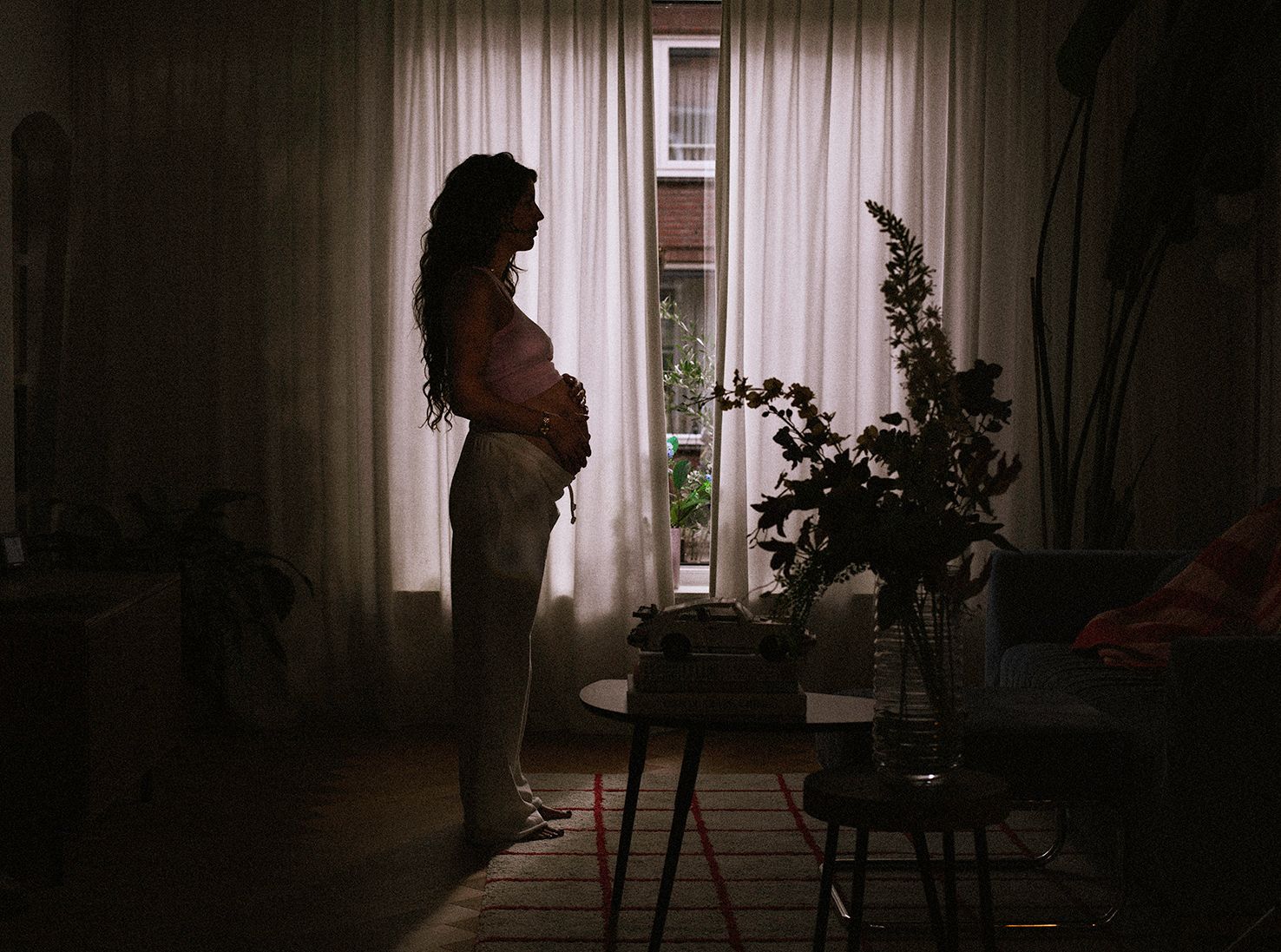 Silhouetted figure by window with dramatic lighting and curtains in dark room.