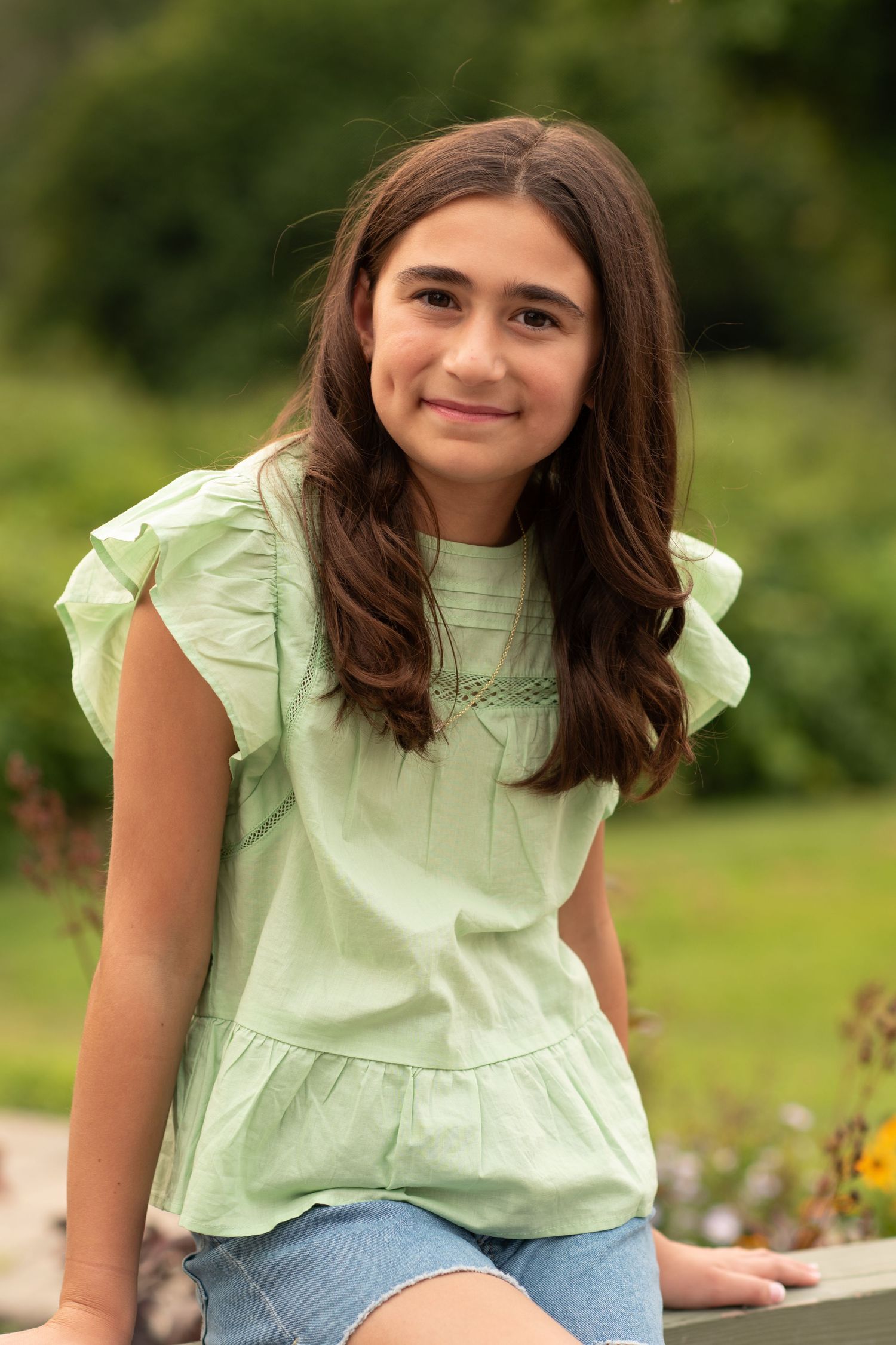Child in green sweater smiling outdoors with autumn foliage in background.