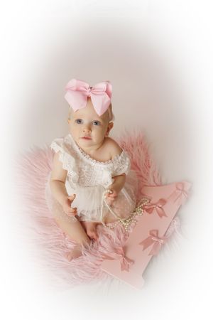Baby girl with a large pink bow sits on a fluffy pink rug in a white lace romper holding a pearl necklace.