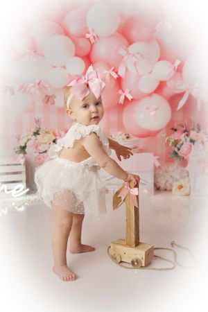 Baby girl in a white tulle romper and pink bow stands holding a wooden push toy in front of a pink balloon backdrop.