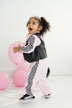 Toddler in a tiara, pink and black racing jumpsuit with checkered stripes and Vans sneakers holds a pink balloon, mouth open in excitement.