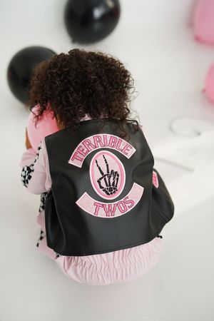 Toddler with curly hair wears a black leather vest with pink Terrible Twos skeleton peace hand patch, sitting among pink and black balloons.