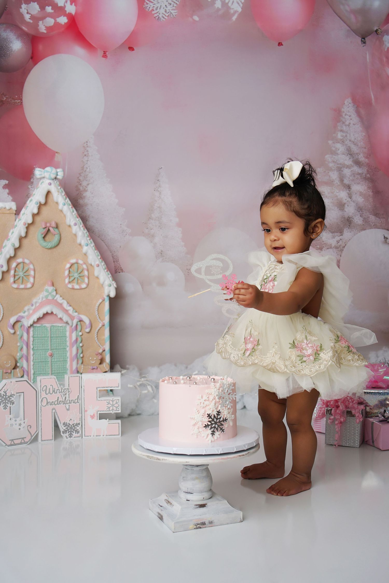 One-year-old girl in a cream tulle dress and bow reaches for a pink snowflake cake in a pink winter wonderland ONE birthday scene.