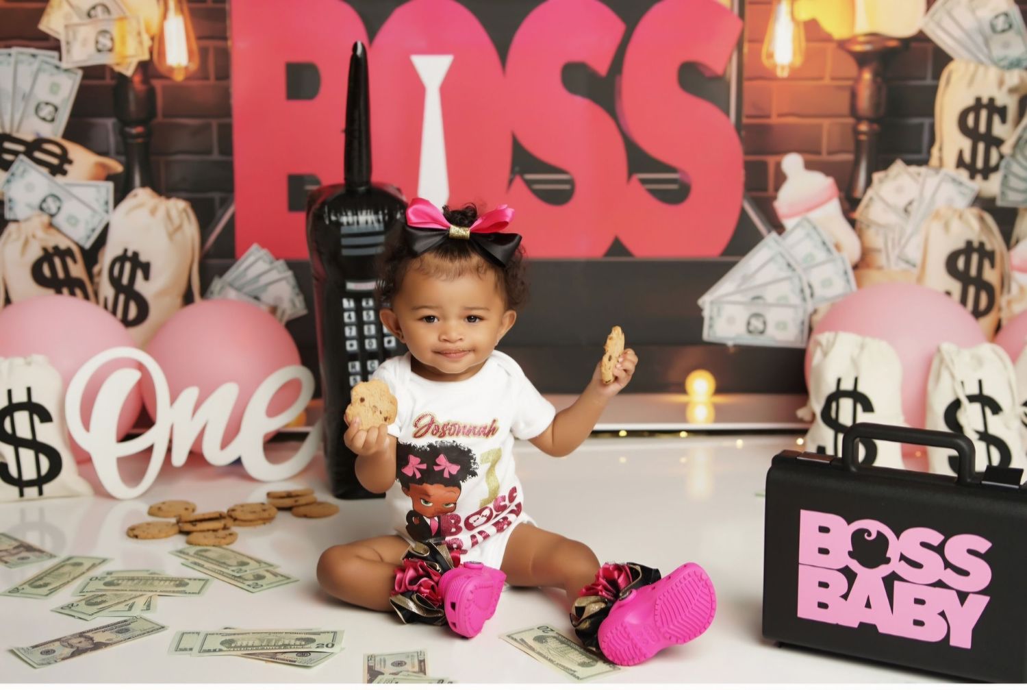 Baby girl in a graphic bodysuit holds a cookie on a first birthday boss-themed set with money bags, balloons and a Boss Baby box.