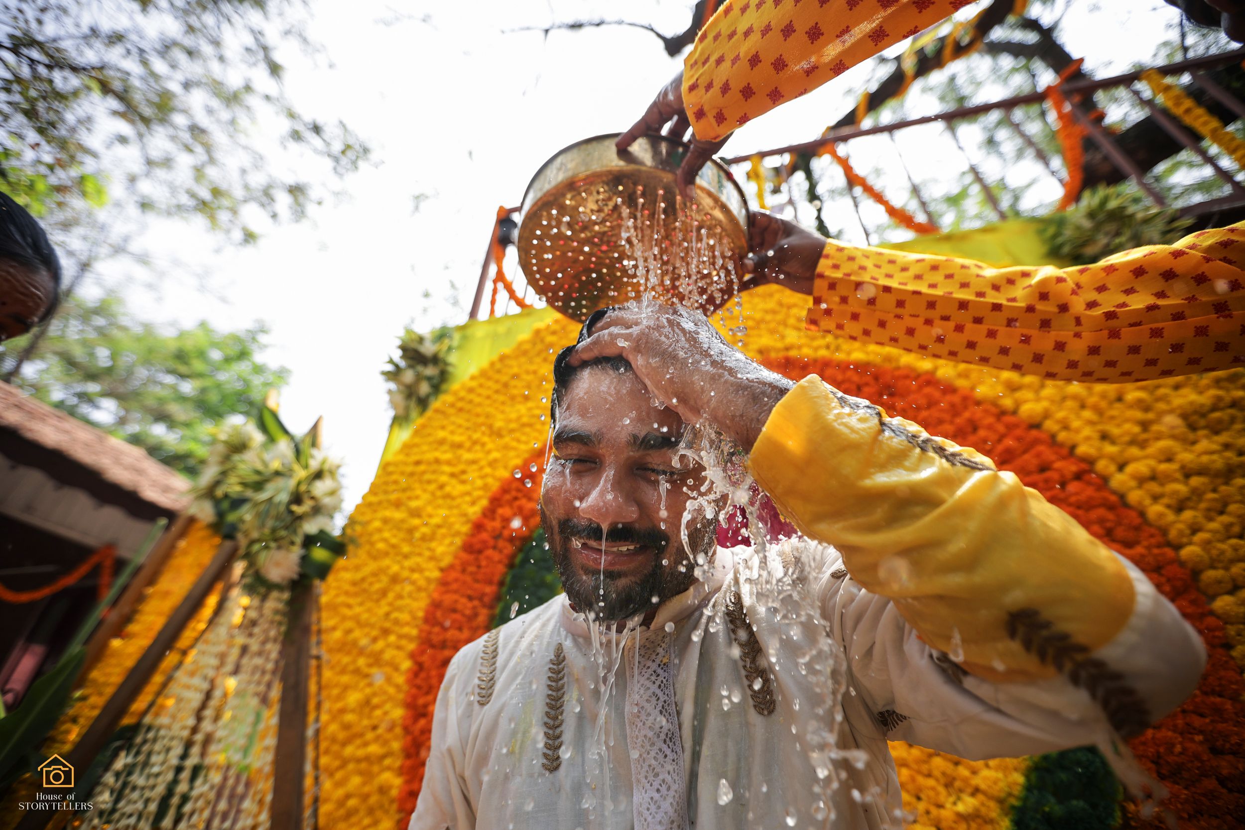 Person performing traditional water ritual under orange and yellow decorative canopy.