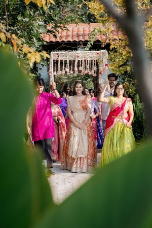 Bride in ivory lehenga walks with bridesmaids in colorful sarees under floral canopy at Indian wedding.