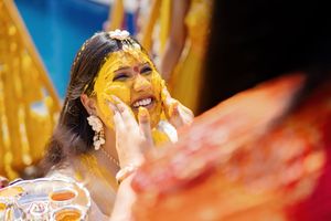 A bride smiles joyfully as turmeric paste is applied to her face during a traditional Haldi ceremony.