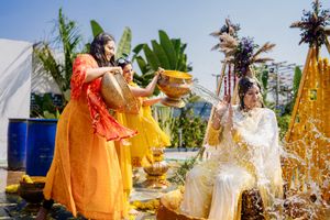 Bride in white outfit gets haldi ceremony with women in yellow pouring water outdoors at Indian wedding.