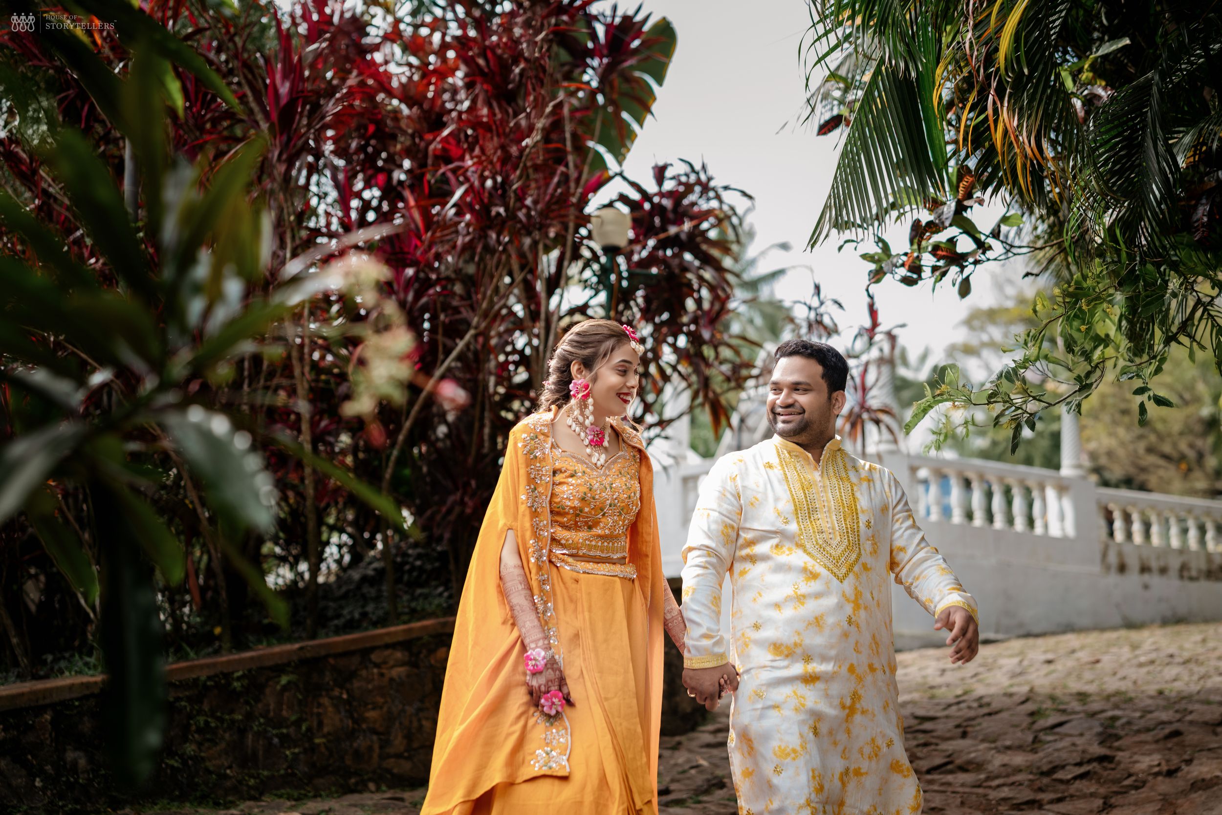 Couple in traditional Indian wedding attire walking through lush tropical garden, woman in yellow lehenga, man in white sherwani.