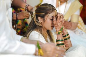A bride in white and gold attire has mehndi-covered hands raised during a traditional Indian wedding ceremony.