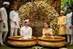 Couple sits in golden tubs at Indian wedding ceremony with floral backdrop and flower petals raining down.