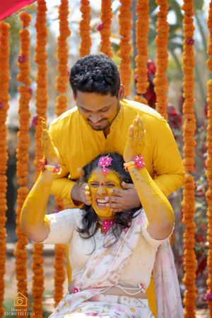 Couple plays with colorful powder and flowers during a traditional Haldi ceremony with marigold garlands in background.