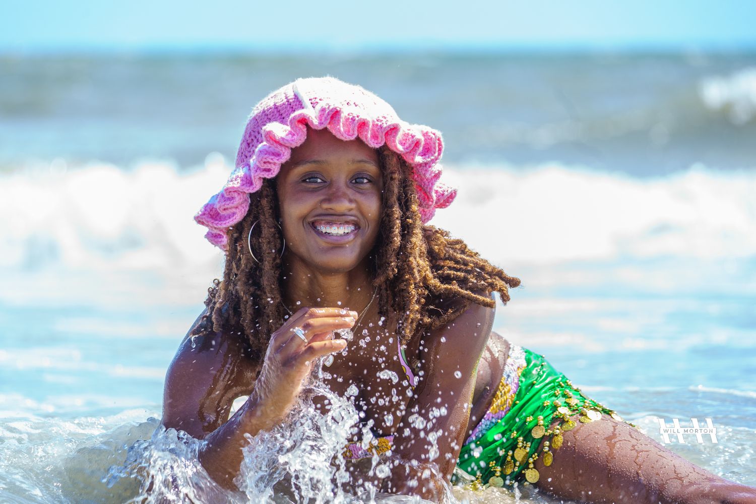 Beachgoer wears pink ruffle hat while playing joyfully in ocean waves.