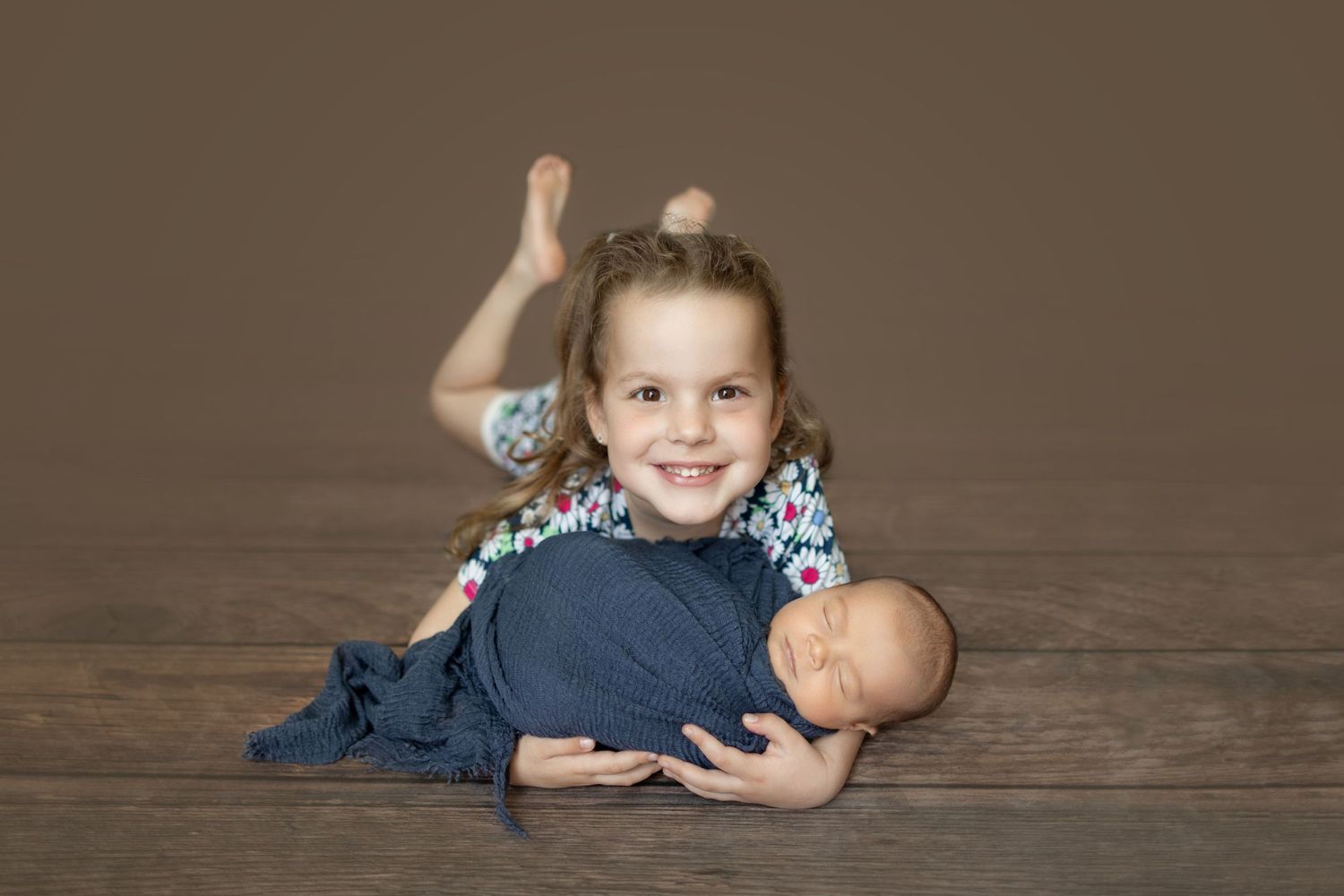 Child lying on wooden floor with a baby doll during a professional photo session.
