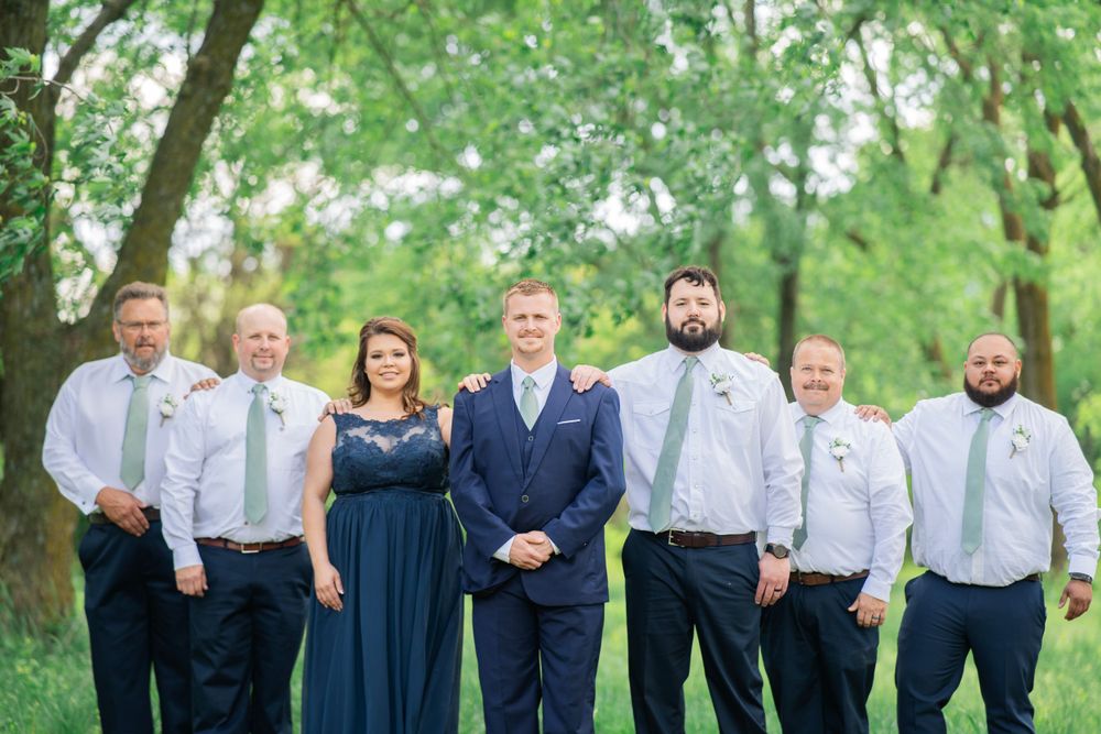 Wedding party in navy and sage green attire posing together under lush green trees.
