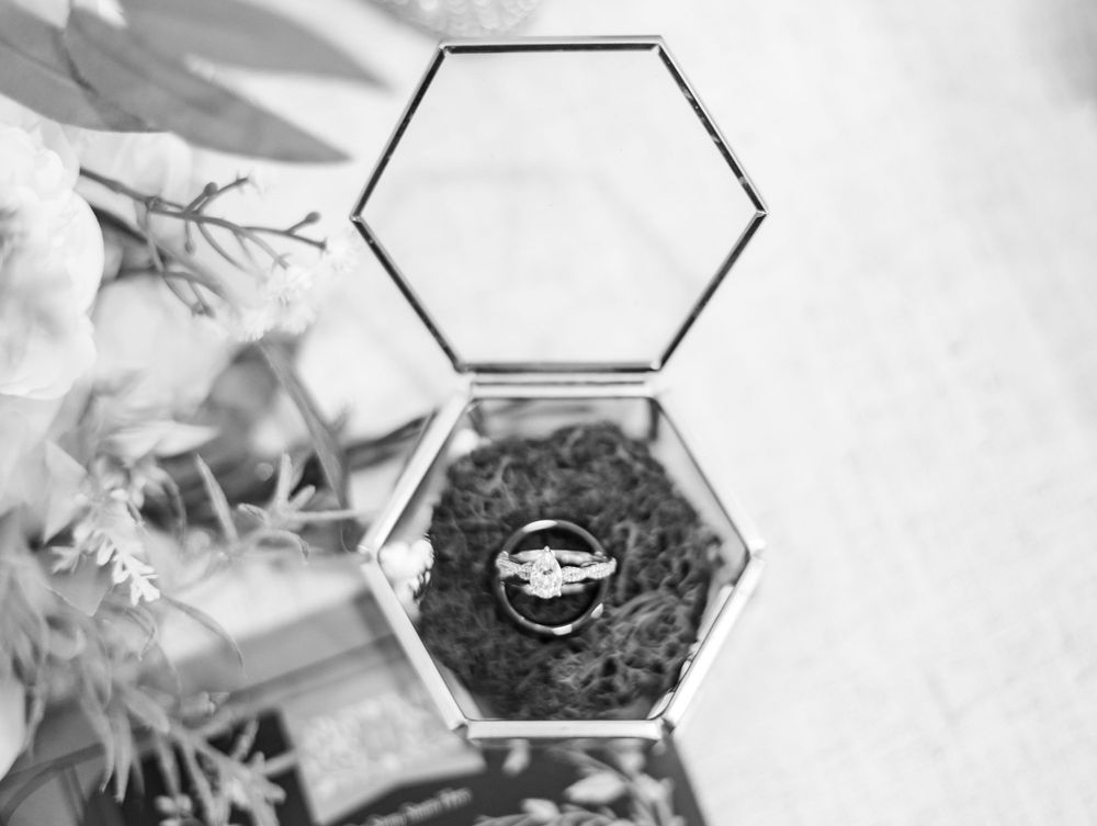 Black and white photo of wedding rings in an open hexagonal glass terrarium box with moss and florals.
