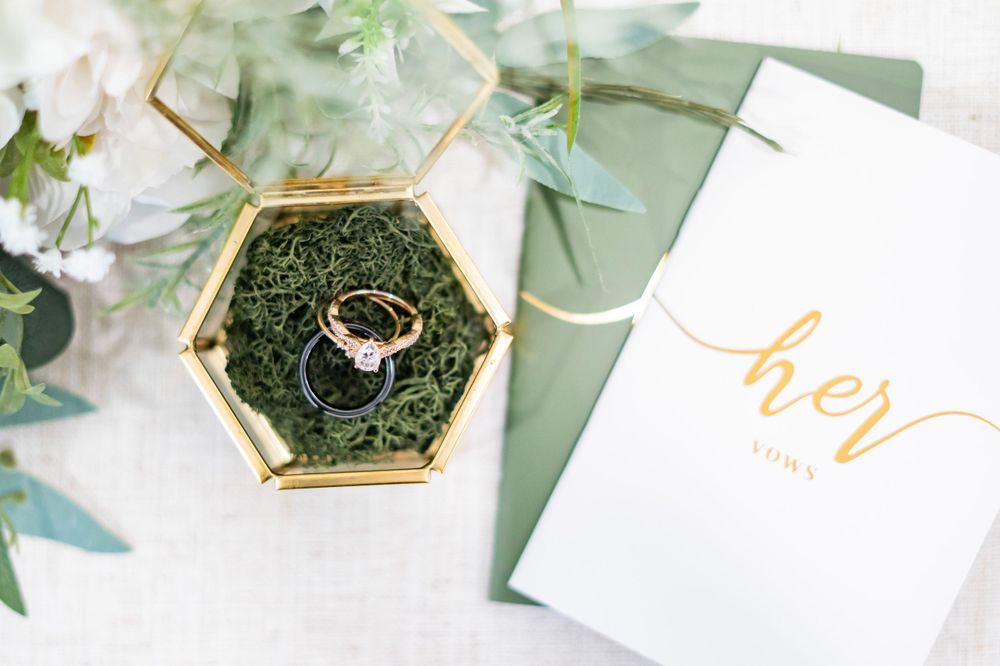 Wedding rings displayed in a gold hexagonal glass terrarium box with moss beside a green and white card.