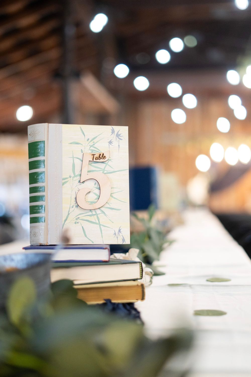 Book-themed wedding table number five displayed on stacked books at a rustic barn reception with string lights.