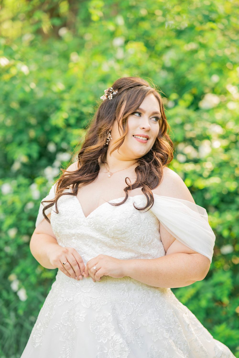 Brunette bride in off-shoulder lace wedding gown with floral hair accessory outdoors.