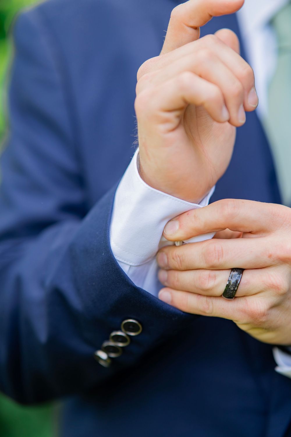 Groom in navy suit adjusting cufflinks, wearing a black wedding band.