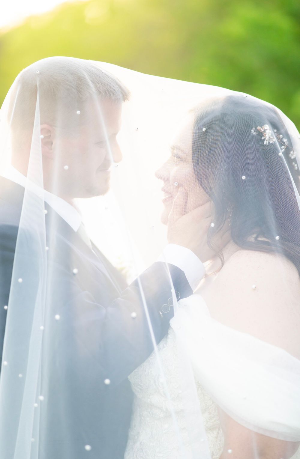 Bride and groom gazing at each other under a pearl-dotted veil with golden sunlight in background.