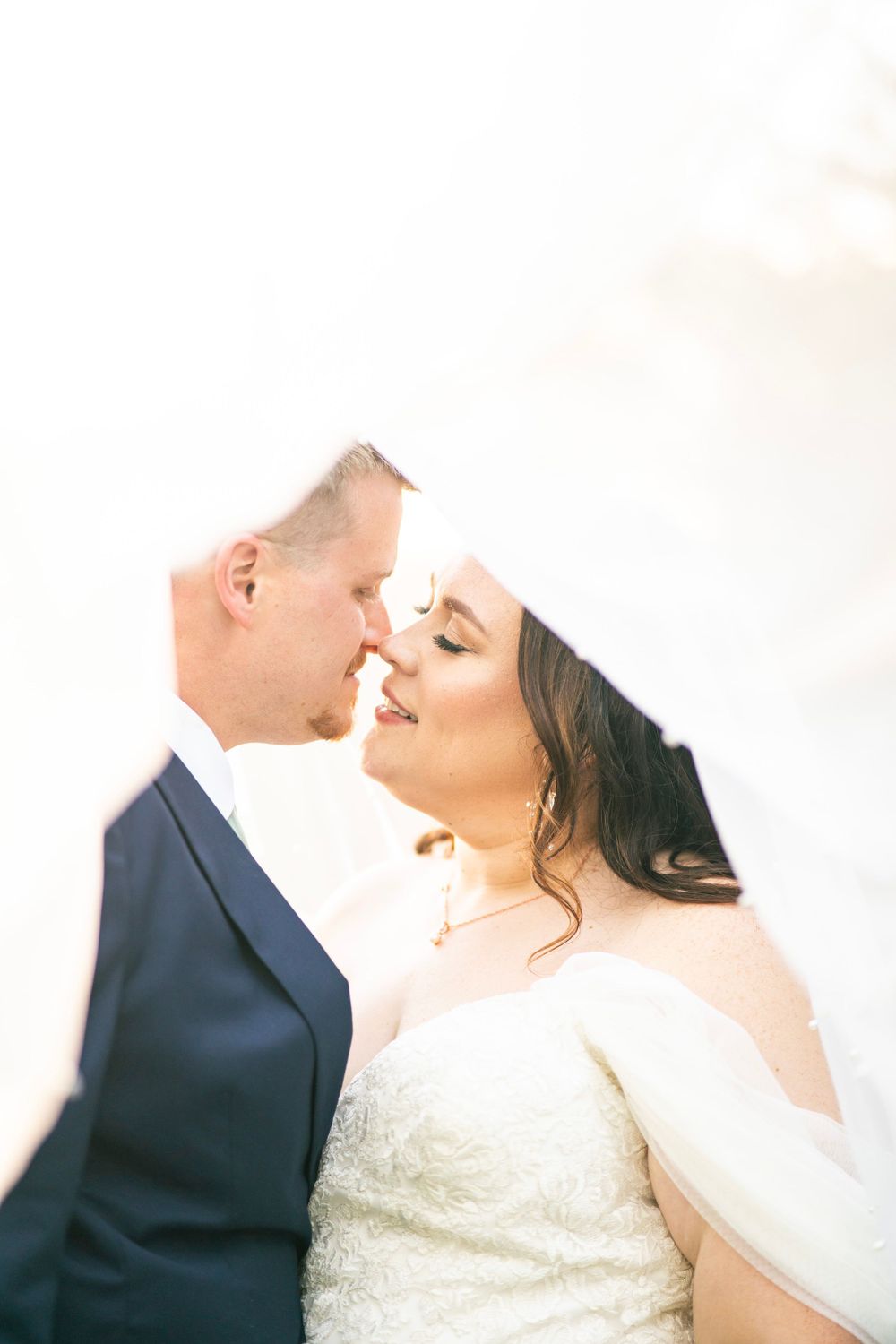 Bride and groom sharing a romantic nose-to-nose moment under a flowing white veil in soft light.