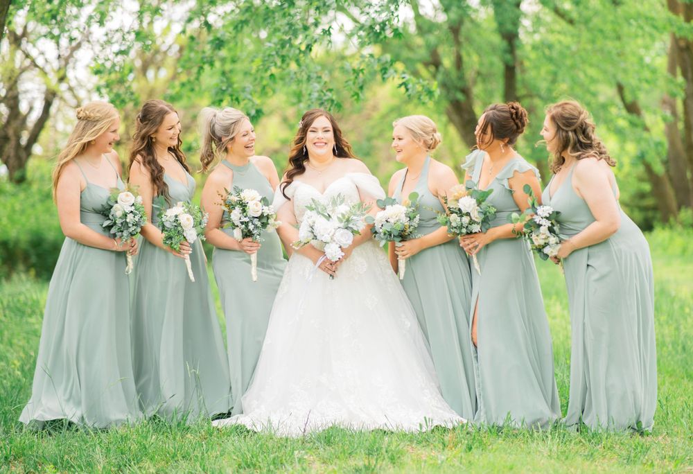 Bride surrounded by six bridesmaids in sage green gowns holding white and green bouquets.