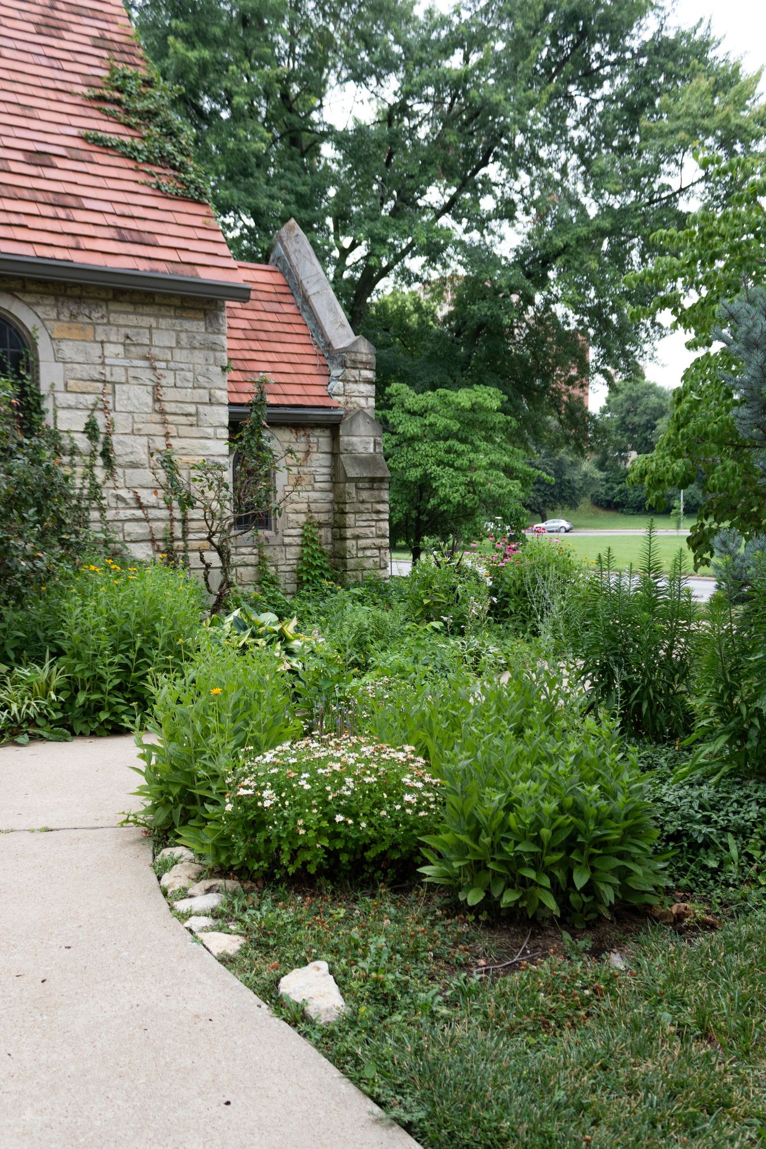 Stone building with red roof surrounded by lush garden pathways and flowering plants.