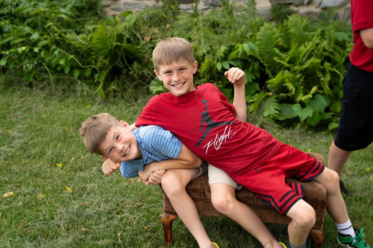 Two children playfully wrestle and laugh on green grass in a garden setting.
