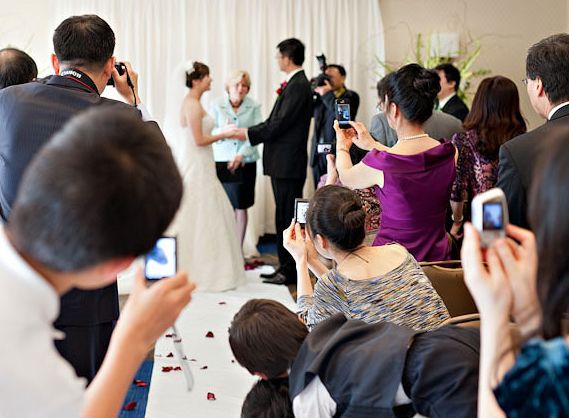 Wedding ceremony guests capturing the moment on smartphones in an indoor venue.