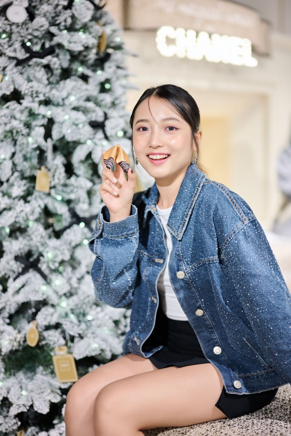 Smiling young Asian woman in denim jacket sits beside a flocked Christmas tree outside a Chanel store holding a small gift.