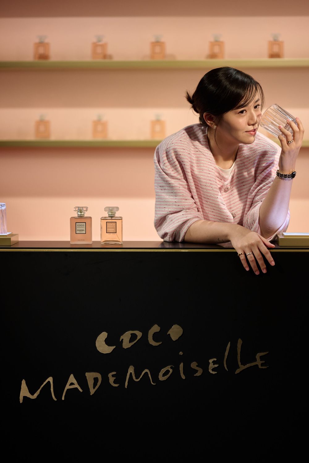 Woman in pink cardigan posing at a Chanel Coco Mademoiselle perfume display counter with bottles on shelves.
