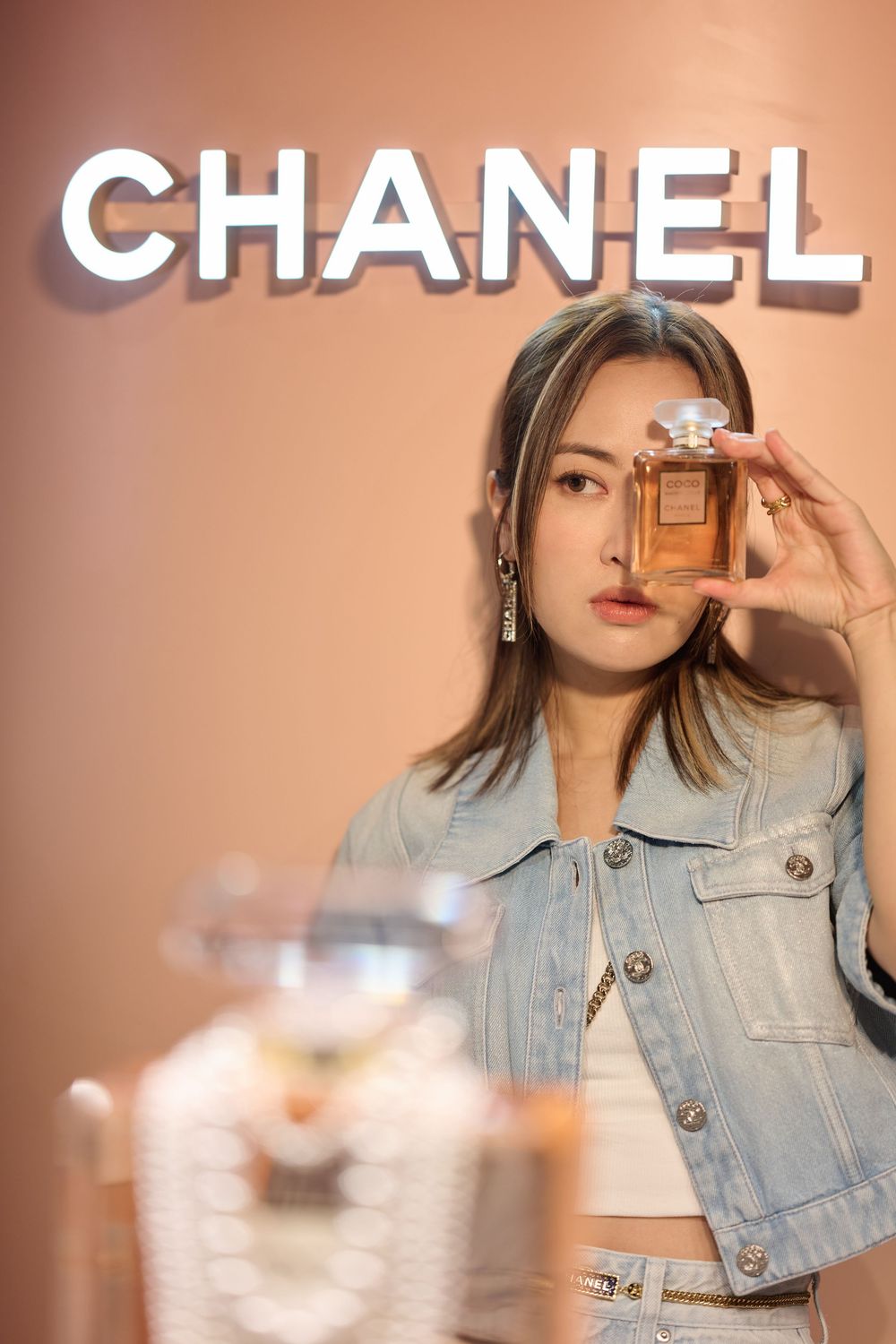 Woman holding Chanel perfume bottle in front of illuminated Chanel sign wearing denim jacket.