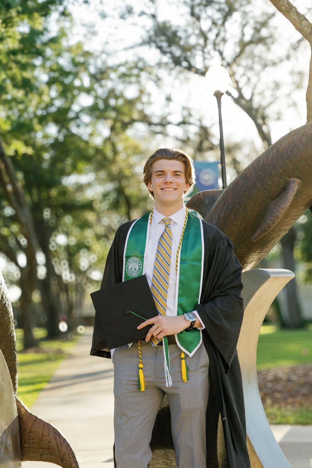 Smiling male graduate in black robe and green stole holding cap at Jacksonville University campus.