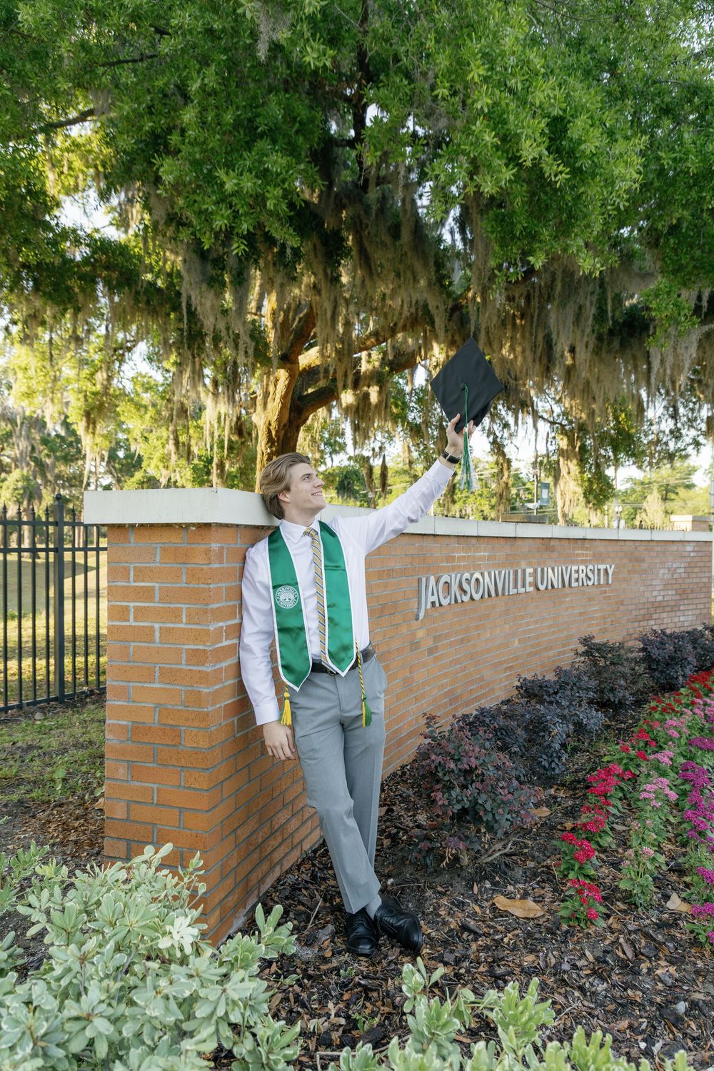 Graduate in academic regalia tossing cap beside Jacksonville University entrance sign outdoors.
