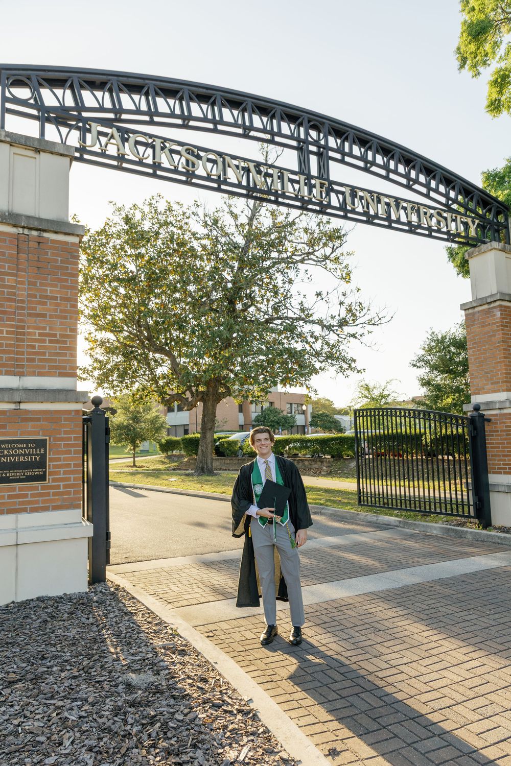 Male graduate in black gown walking through Jacksonville University ornate iron entrance gate.