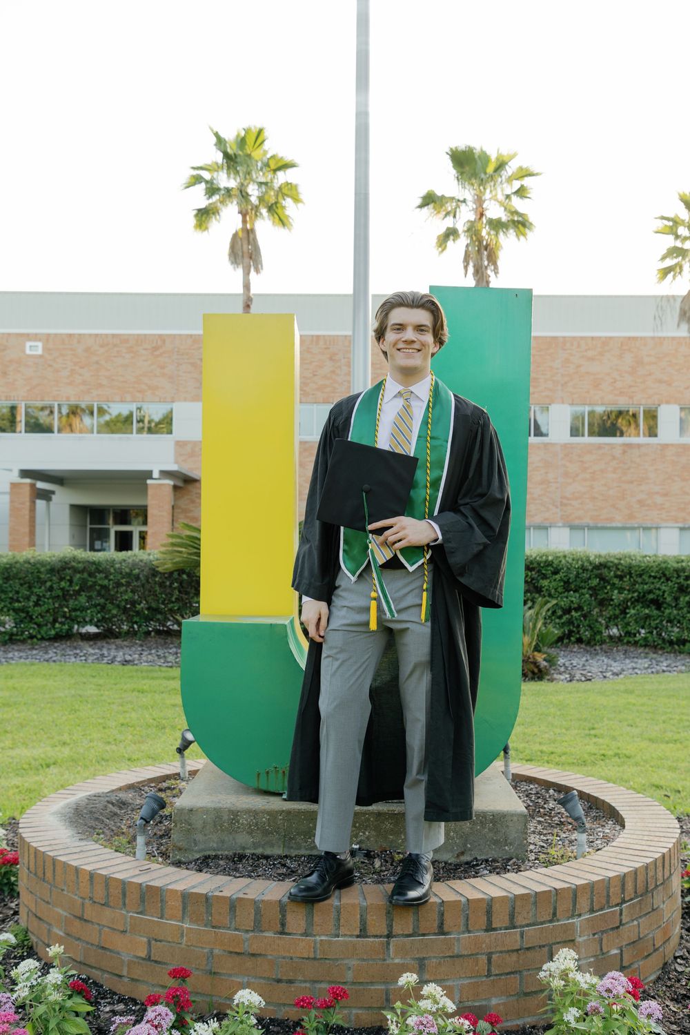 Proud graduate in black robe posing before large green and yellow JU letters on campus grounds.