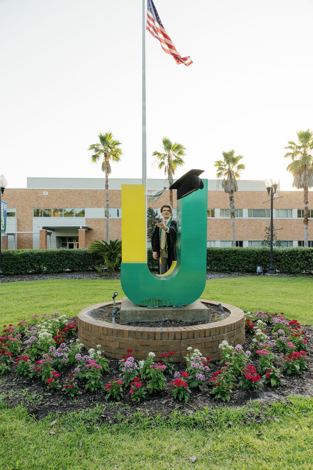 Graduate in cap and gown posing inside large green JU letter sculpture with American flag backdrop.