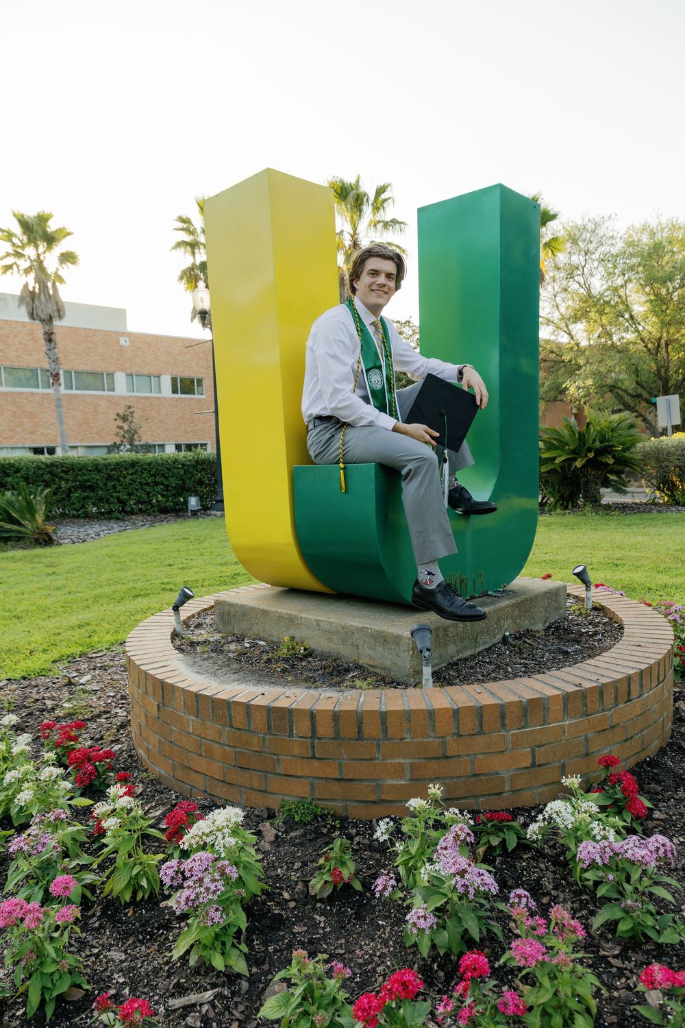 Graduate sits in large green and yellow JU-shaped university sculpture surrounded by colorful flowers.