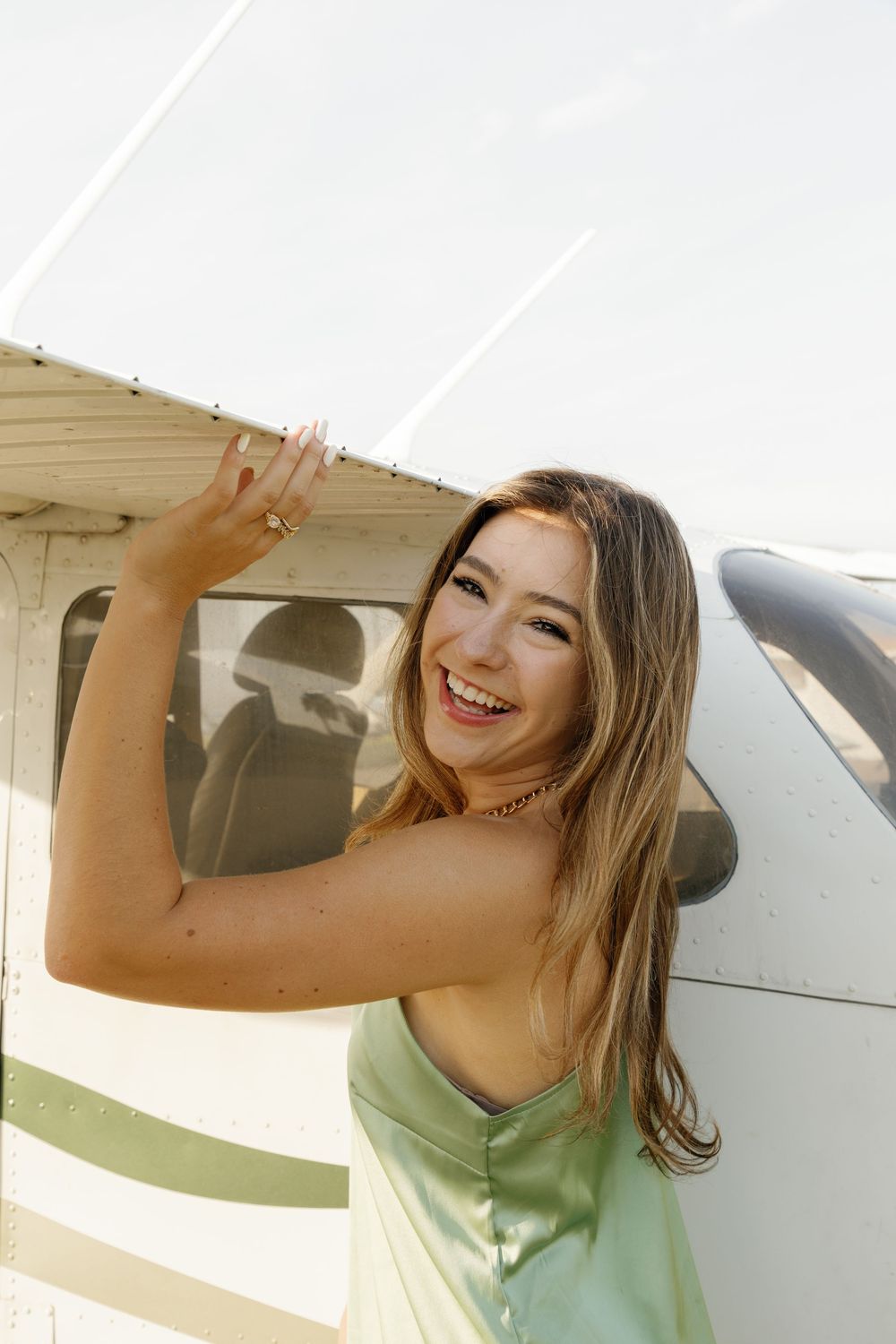 Smiling young woman in green dress posing beside a small white aircraft at an airport.
