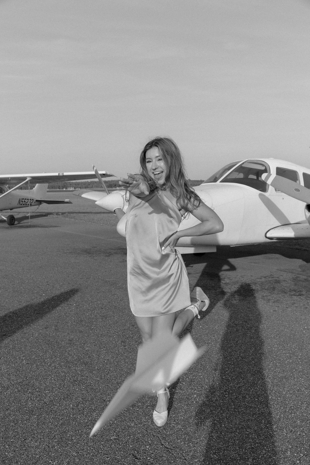 Black and white photo of joyful young woman in dress posing with small aircraft on airport tarmac.