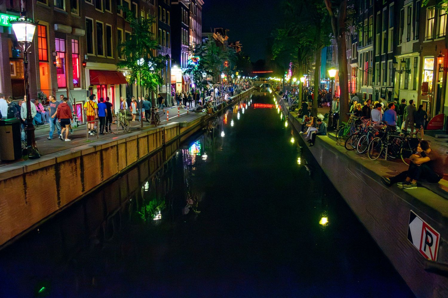 Amsterdam canal at night with colorful lights reflecting on dark water and busy sidewalks.