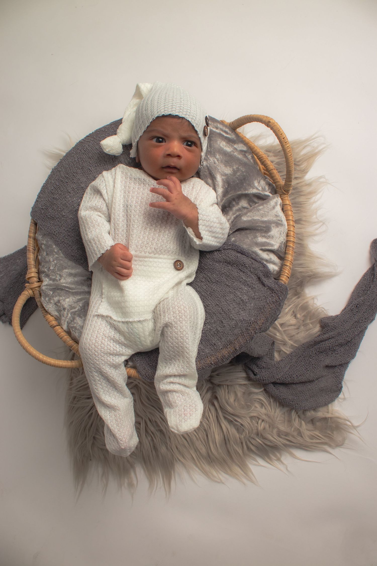 Baby wearing white knitted outfit lying on grey furry blanket in basket.