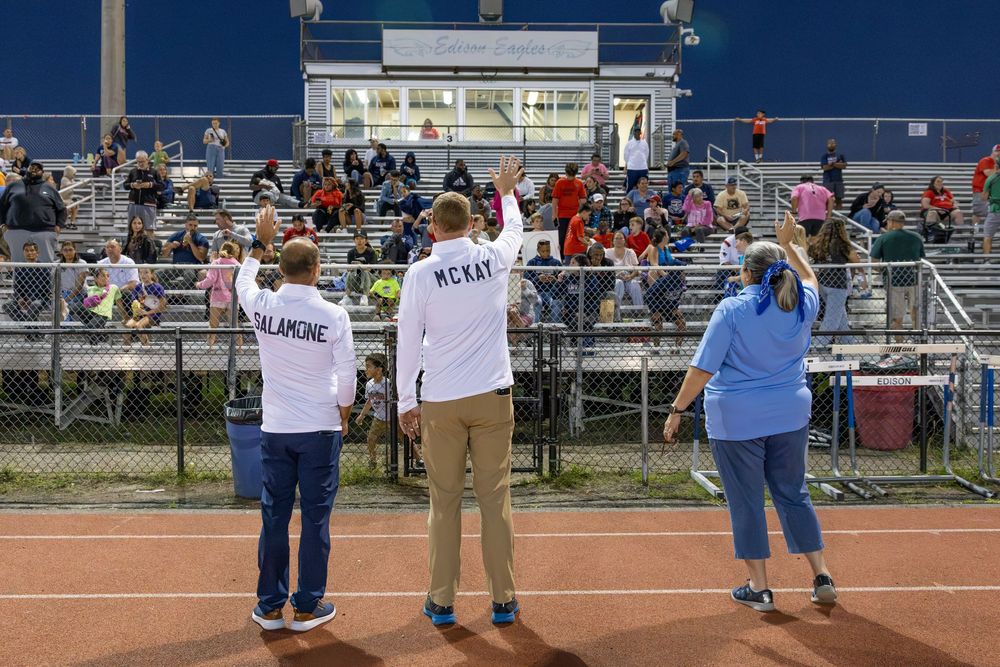 Three people wearing face masks stand socially distanced on a baseball field while raising their arms during an outdoor event.